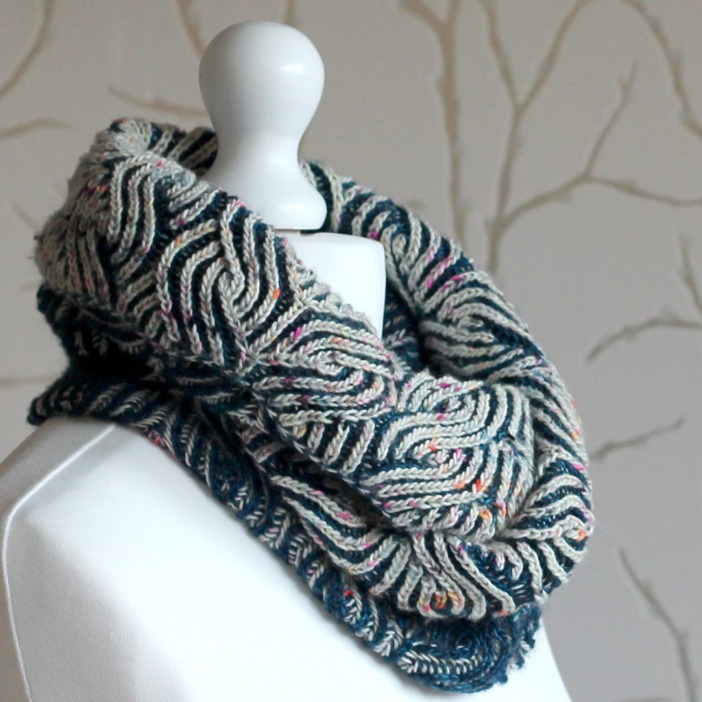 A mannequin displaying a long brioche cowl in blue and white with a owl pattern wrapped twice around the neck