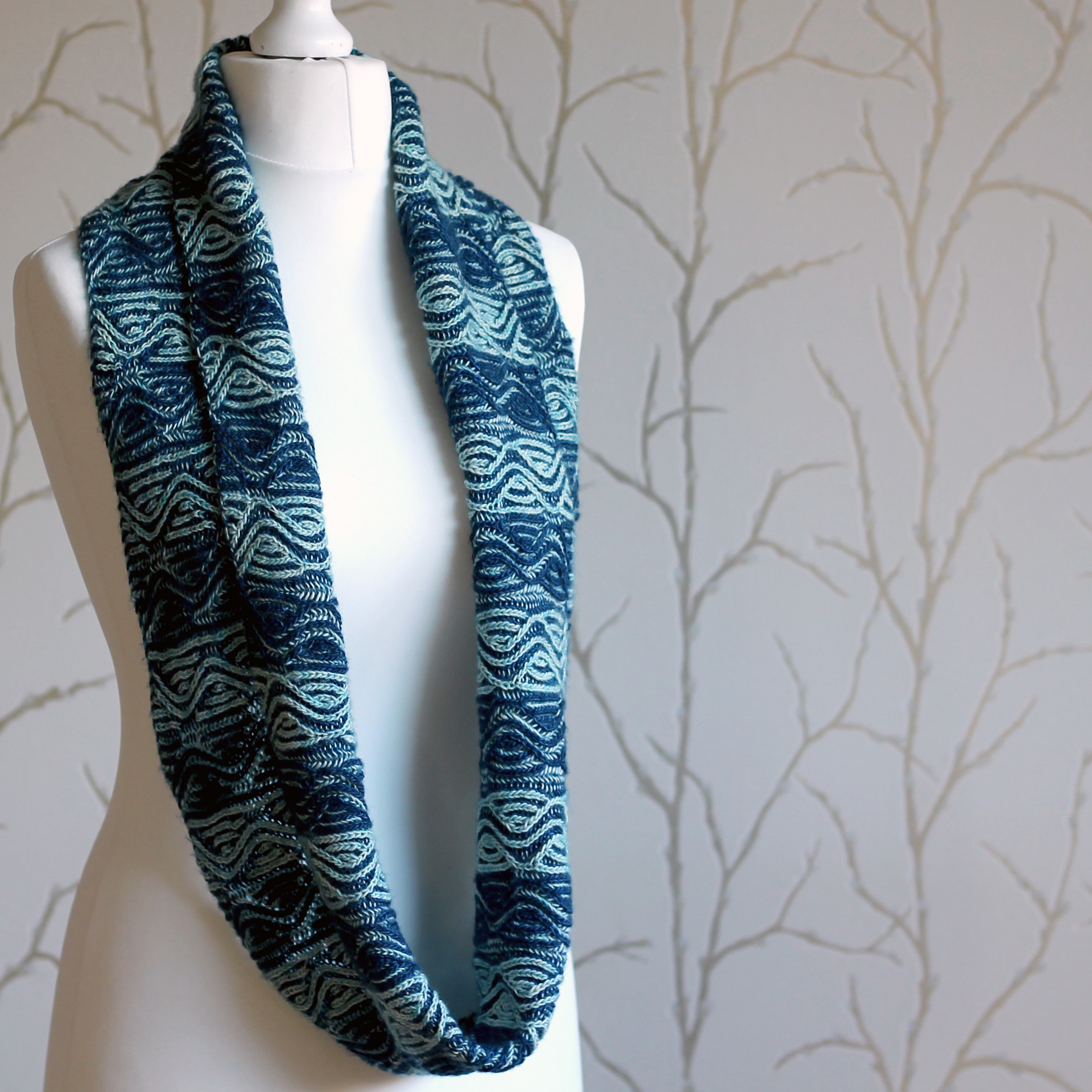 A mannequin displaying a long brioche cowl with dark and light blue vertical stripes and a faux cable pattern
