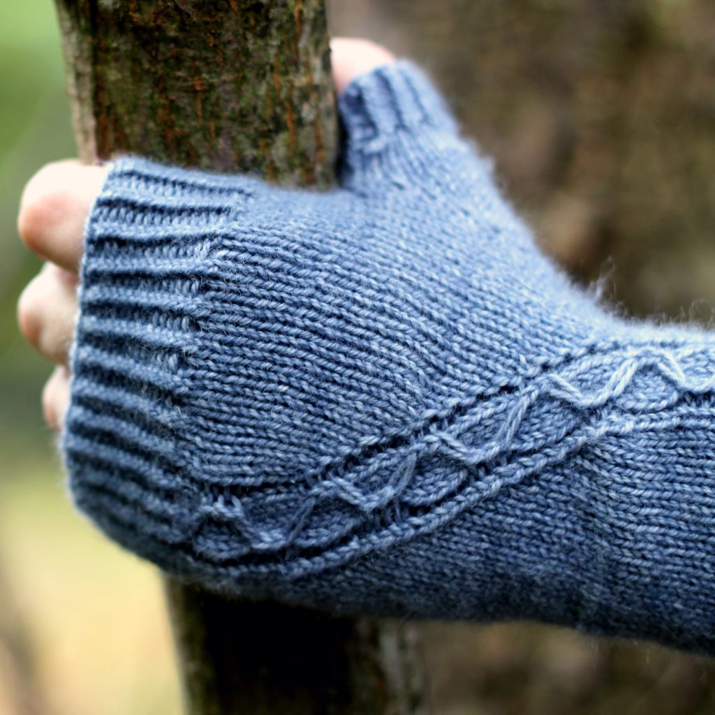 A blue fingerless mitt with a cable pattern travelling diagonally across the back of the hand, with the hand holding a tree branch